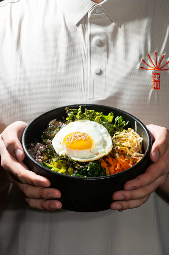A staff member in a white shirt embroidered with the dandelion logo holds a bowl of bibimbap topped with a fried egg