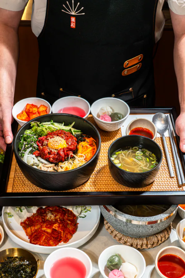 A staff member holds a rattan tray with Hanwoo beef tartare bibimbap in a black bowl, soup and side dishes